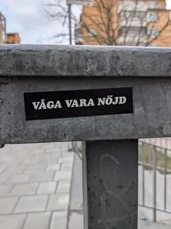 A rectangular black sticker with bold white uppercase text, reading 'VÅGA VARA NÖJD'. The sticker is applied horizontally to a weathered, grey metal beam, showing slight wear and tear around its edges. The text uses a clear, sans-serif font. The overall aesthetic is minimalist and direct.