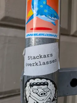 A white sticker with a slightly wrinkled texture, affixed to a metal pole. The sticker has black text printed on it in a serif font. Below it, another black and white sticker with a cartoonish, somewhat menacing creature can be seen.
