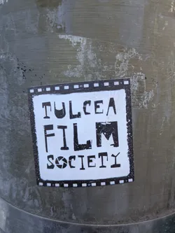 This sticker features a black and white design with the text "Tulcea Film Society". The text is arranged in a stacked format inside a black and white border resembling a film strip. A graphic resembling a camera aperture is placed between the words "Film" and "Society". The sticker seems weathered.