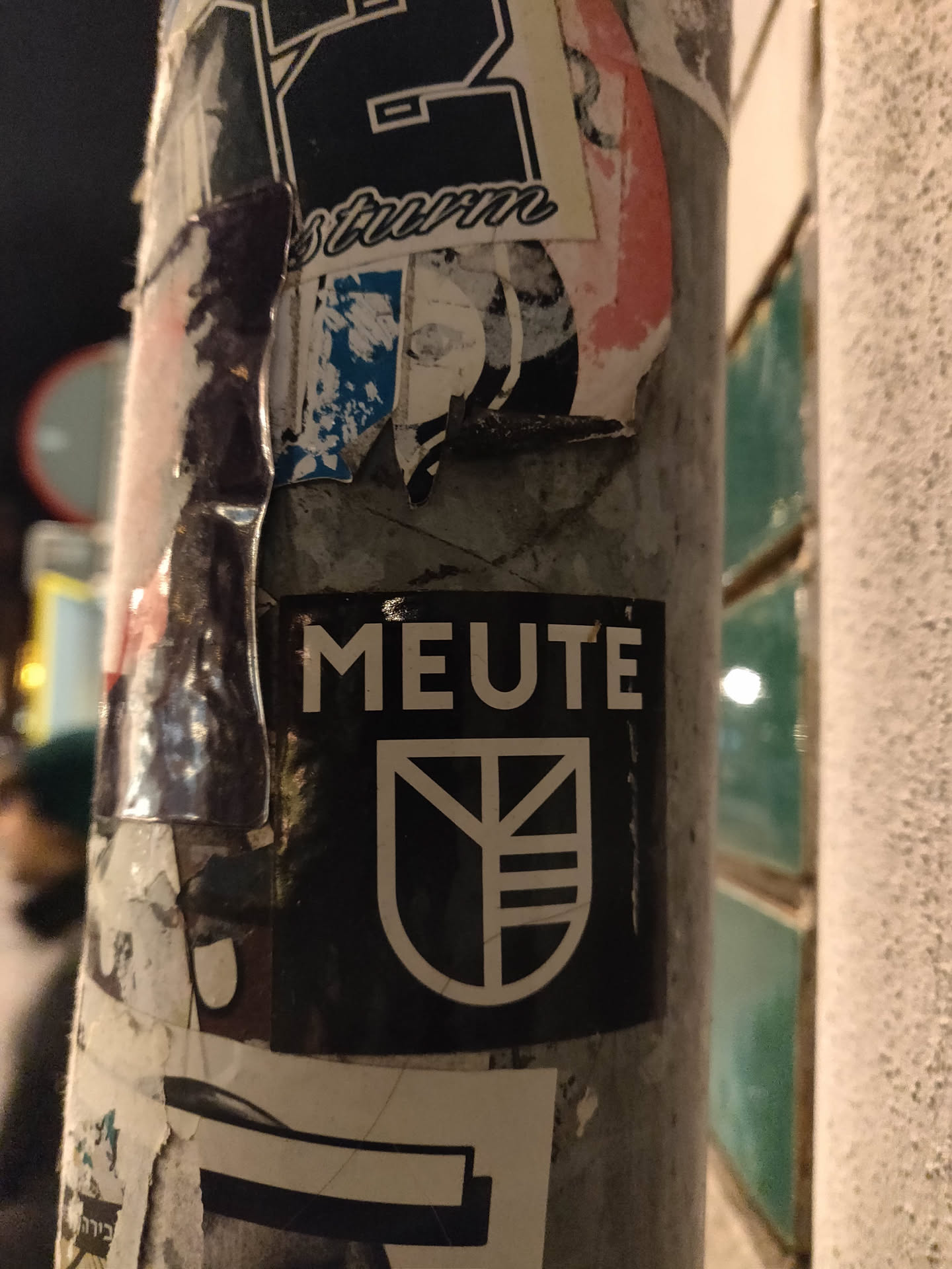 Black and white sticker with the word MEUTE and a logo that looks like a crest. The sticker is on a pole covered in other stickers.