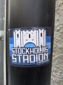 The sticker prominently displays a white, stylized castle with blue outlines and accents, representing a stadium. Below the castle, the text 'STOCKHOLMS STADION' is rendered in large, blocky letters with a metallic silver sheen and blue outlines. In the lower right corner, a smaller, cursive text reads 'Djurgården'. The background of the sticker features horizontal bands of blue and black.