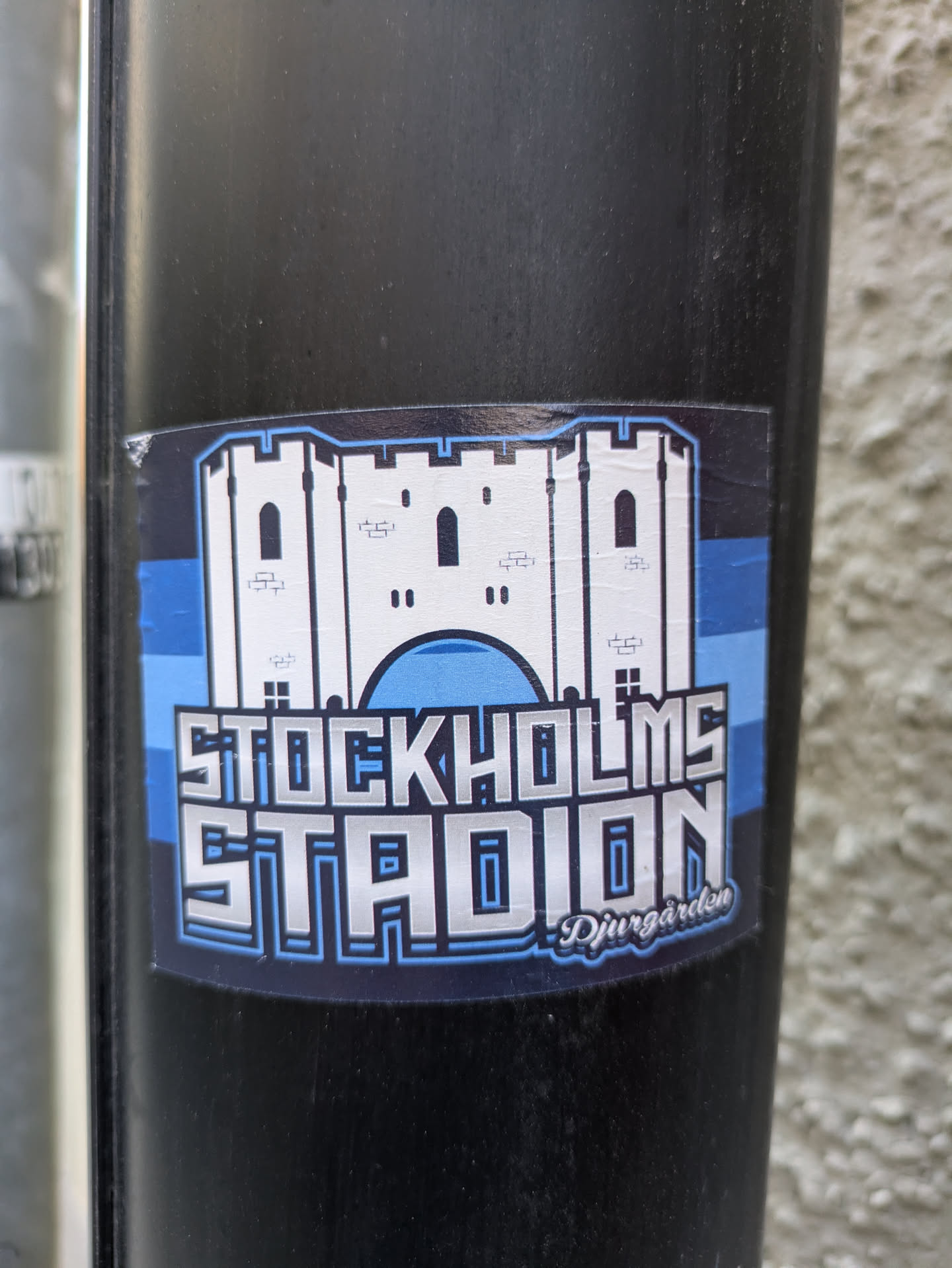 The sticker prominently displays a white, stylized castle with blue outlines and accents, representing a stadium. Below the castle, the text 'STOCKHOLMS STADION' is rendered in large, blocky letters with a metallic silver sheen and blue outlines. In the lower right corner, a smaller, cursive text reads 'Djurgården'. The background of the sticker features horizontal bands of blue and black.