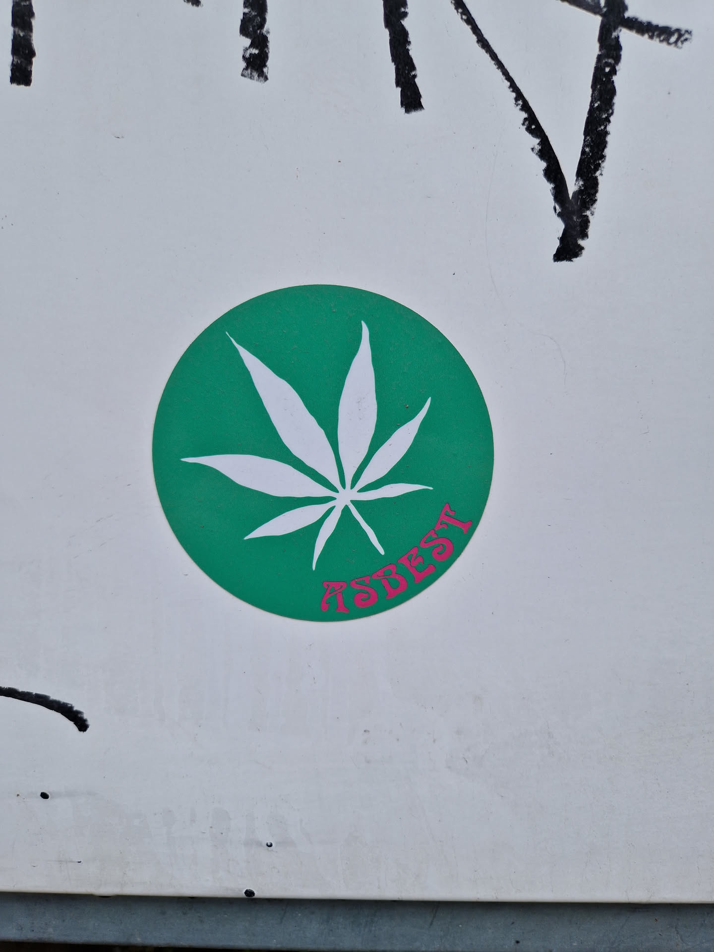 The sticker features a stylized marijuana leaf on a green circular background.  The word "ASBEST" is written underneath the leaf in a pink, stylized font. The sticker is affixed to a white surface with some markings or graffiti visible in the background.