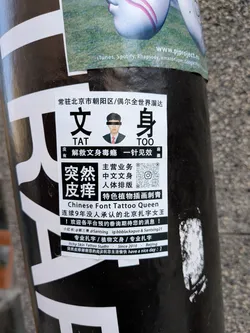 A rectangular white sticker with prominent black Chinese characters and English text, adhered to a black pole. The sticker features a small passport-style photo of a man with a black bar over his eyes. Large characters '文身' (Tattoo) and '突然皮痒' (Suddenly Itchy Skin) are central. It lists various tattoo services, contact information, social media handles, and includes a QR code. The overall design is a mix of professional advertisement and quirky humor, showing slight wear and curvature from being on the pole.