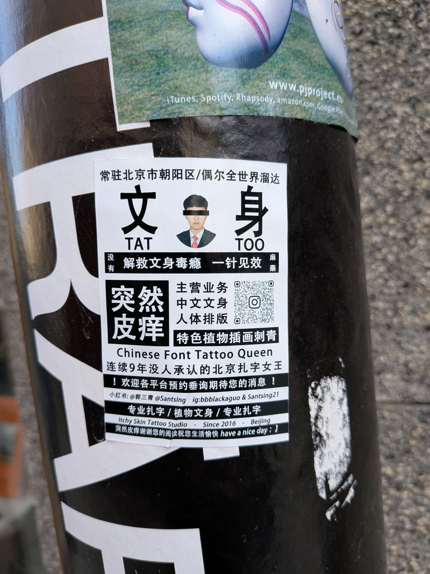 A rectangular white sticker with prominent black Chinese characters and English text, adhered to a black pole. The sticker features a small passport-style photo of a man with a black bar over his eyes. Large characters '文身' (Tattoo) and '突然皮痒' (Suddenly Itchy Skin) are central. It lists various tattoo services, contact information, social media handles, and includes a QR code. The overall design is a mix of professional advertisement and quirky humor, showing slight wear and curvature from being on the pole.