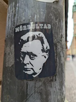 Black and white sticker featuring a portrait of a man with the word "MÖRBULTAD" written above. The sticker is affixed to a weathered pole.