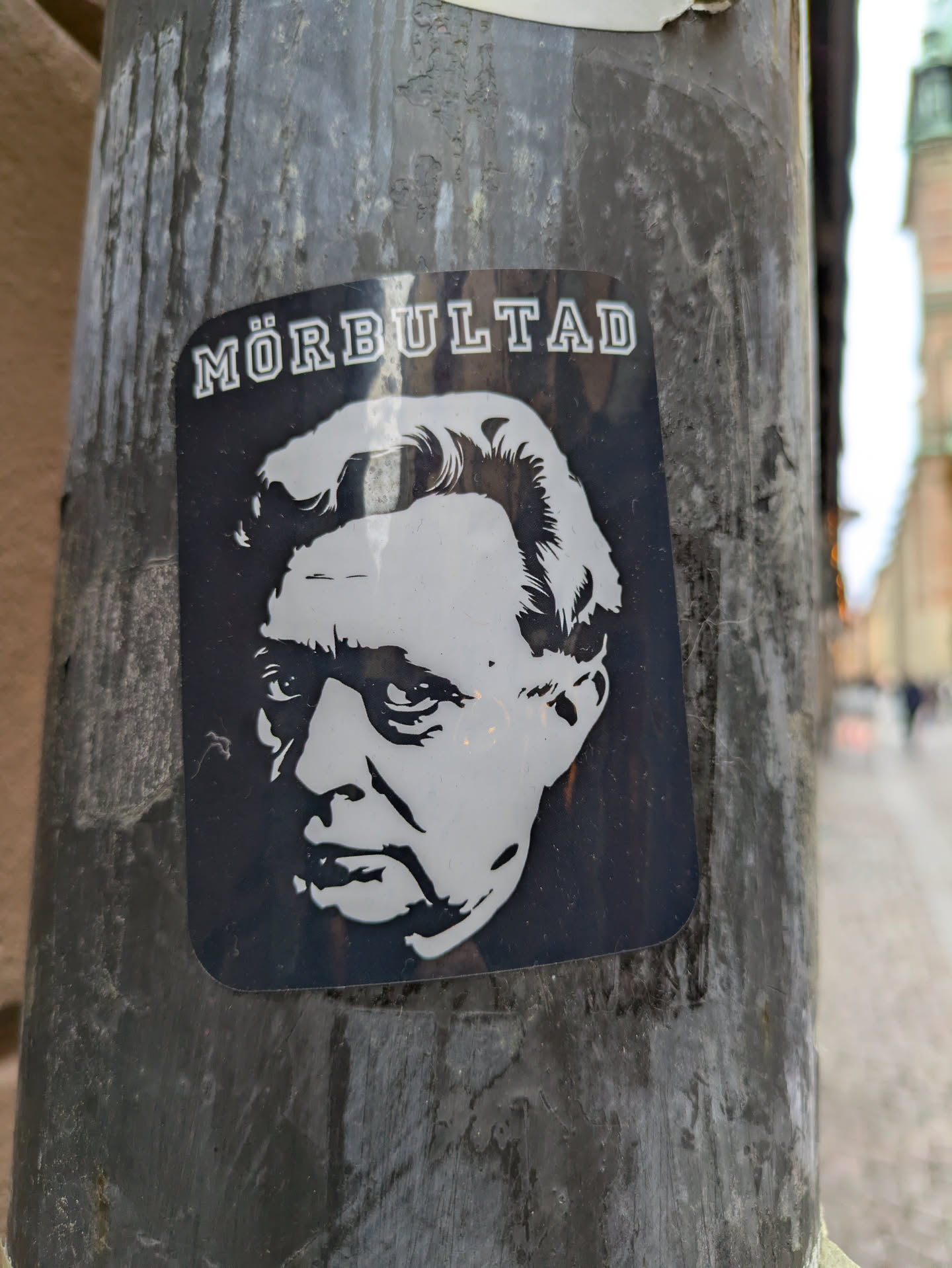 Black and white sticker featuring a portrait of a man with the word "MÖRBULTAD" written above. The sticker is affixed to a weathered pole.