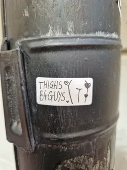 A sticker with the text 'THIGHS ♡ B4 GUYS. T ♡' on a dark-colored metal surface. The sticker has a handwritten style and includes small drawings of hearts and flowers.