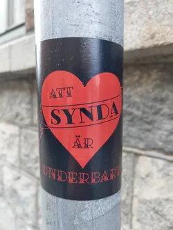 A black and red sticker with a heart shape.  The text on the sticker says "Att synda är underbart" which translates from Swedish to English as "To sin is wonderful".