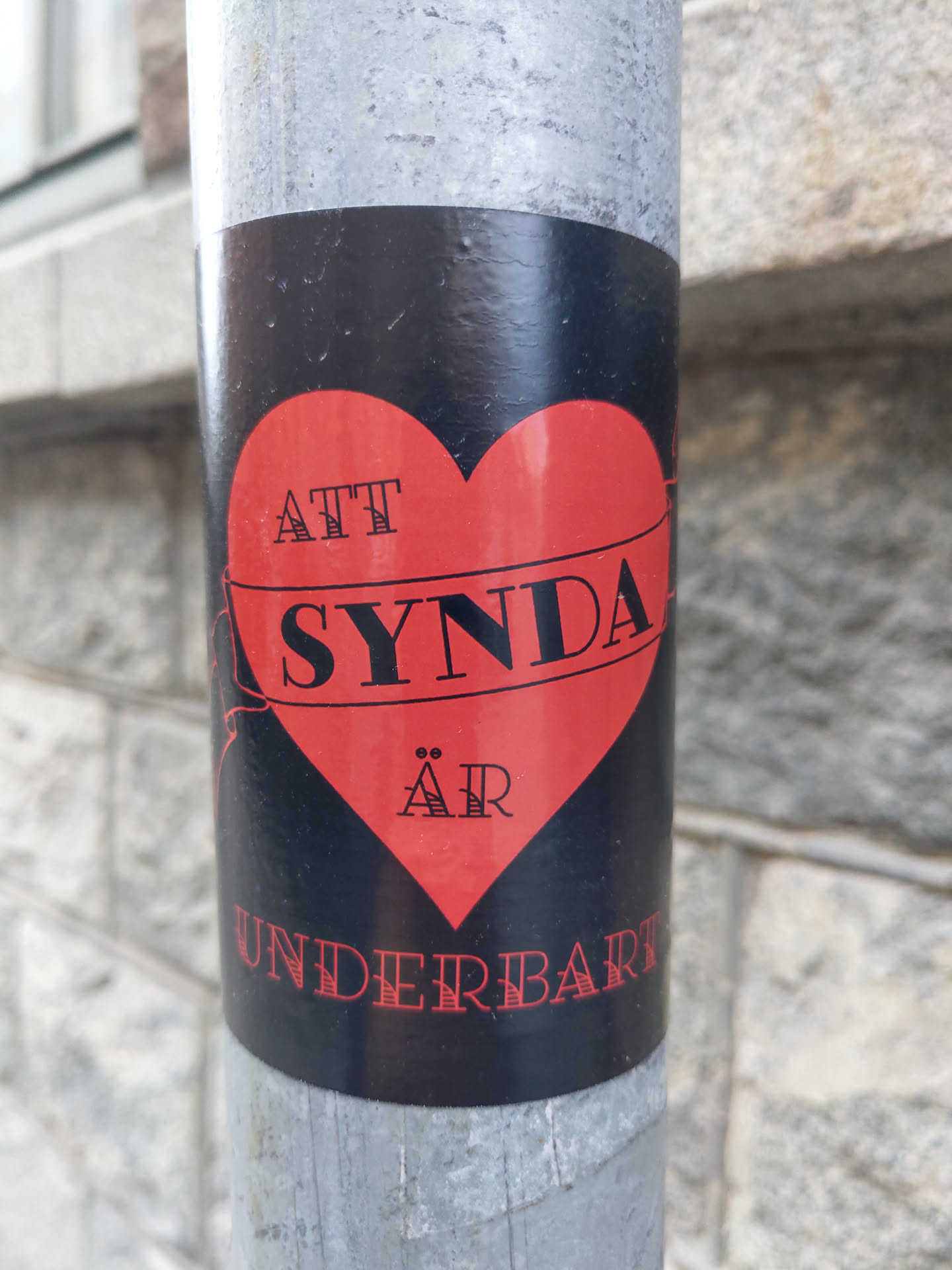 A black and red sticker with a heart shape.  The text on the sticker says "Att synda är underbart" which translates from Swedish to English as "To sin is wonderful".