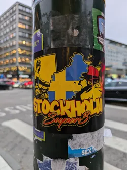 A sticker featuring a stylized graphic of a map of Sweden, possibly representing Stockholm, along with illustrations of a muscular man and a figure in a cap. The text "STOCKHOLM Super 3" is prominently displayed.
