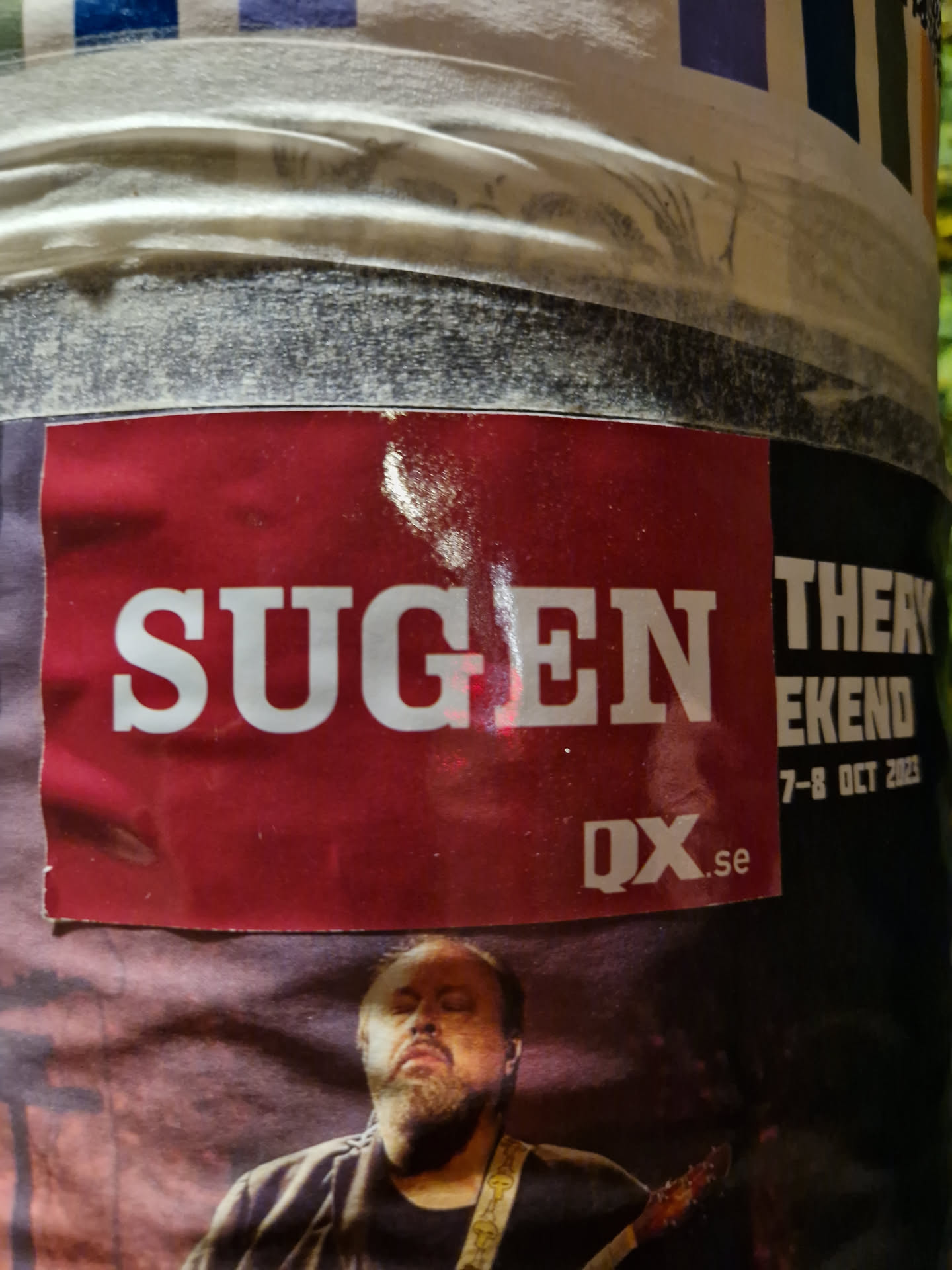 A red sticker with the word SUGGEN in white capital letters.  The website QX.se is visible in the lower right corner. Part of a larger poster advertising a music event (partially visible text includes 'Weekend', '7-8 Oct 2023'). A picture of a man, possibly a musician, is visible underneath.