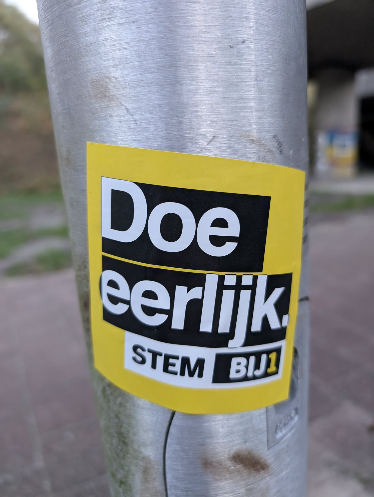 The sticker is predominantly yellow with black and white text. The top portion features the word 'Doe' in large white sans-serif font against a black background. Below this, separated by a thin yellow line, is the word 'eerlijk.' also in white sans-serif font on a black background. At the bottom, within a white rectangular box with a black outline, are the words 'STEM BIJ1' in black text. The yellow background extends beyond the black text areas, giving the sticker a dynamic, slightly angled appearance.