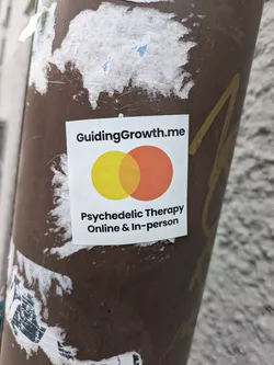 A sticker advertising psychedelic therapy services, both online and in-person, from Guiding Growth. The sticker features a logo with two overlapping circles in yellow and orange. 