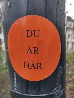 Orange round sticker on a dark pole with the text "DU ÄR HÄR" which translates to "You are here" in Swedish.