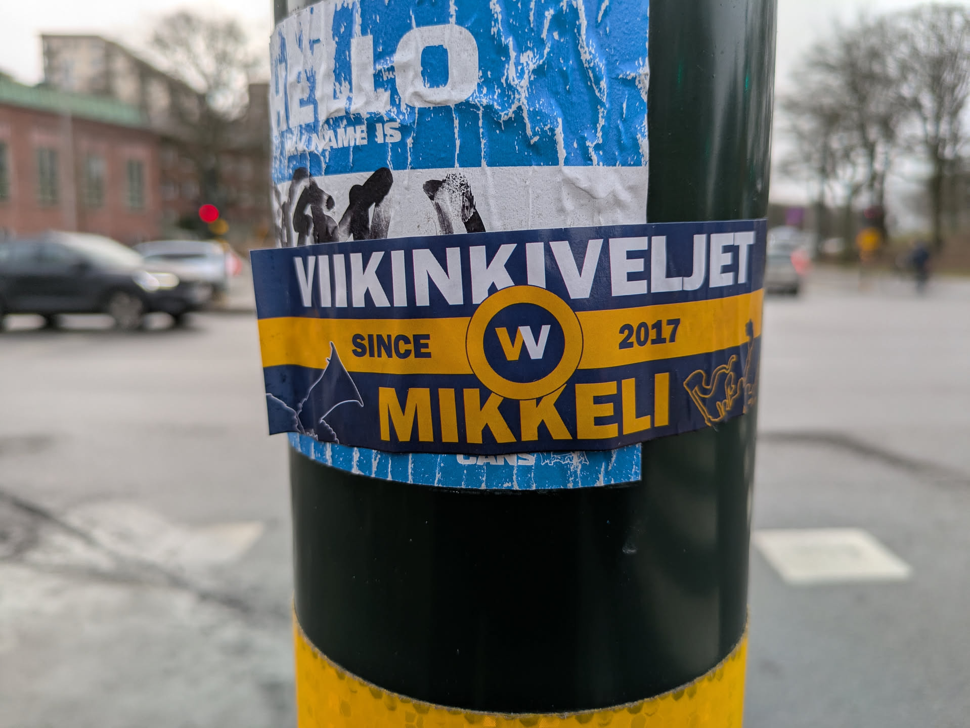 The image shows a Viikinkiveljet Mikkeli sticker on a pole. The sticker is mostly blue and yellow. The text on the sticker reads "VIIKINKIVELJET SINCE 2017 MIKKELI".