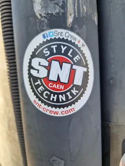 Round sticker with a red and black design. It has the text 'STYLE' on top, 'SNT' in large letters in the center, 'CAEN' under 'SNT', and 'TECHNIK' below 'CAEN'. At the bottom, it says 'snt-crew.com'. Social media links (Facebook, Twitter, Instagram) are mentioned above the main logo.