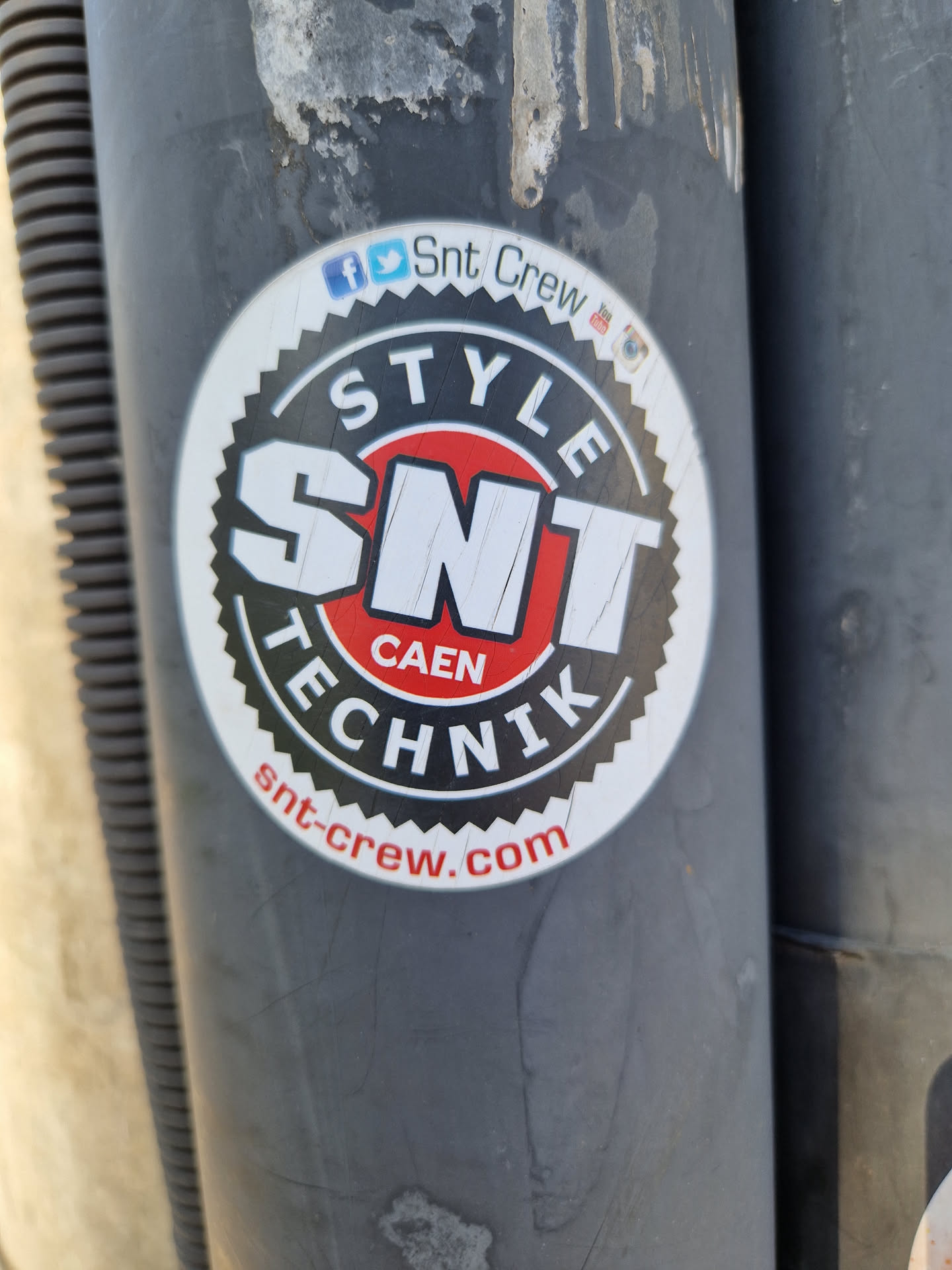 Round sticker with a red and black design. It has the text 'STYLE' on top, 'SNT' in large letters in the center, 'CAEN' under 'SNT', and 'TECHNIK' below 'CAEN'. At the bottom, it says 'snt-crew.com'. Social media links (Facebook, Twitter, Instagram) are mentioned above the main logo.