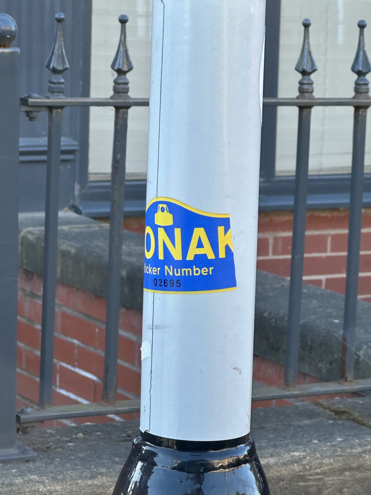 The sticker is predominantly royal blue with bright yellow text and a yellow icon, outlined by a thin yellow border. Its shape is generally rectangular with a wavy, slightly curved top edge. The most prominent feature is the large, bold, yellow capital letters 'MONAK' in the center. Above the 'O' in 'MONAK', there is a yellow icon resembling a bell or a small bag/purse with a rounded top and a wider base. Below 'MONAK', in smaller yellow text, it reads 'Sticker Number', followed by the numerical identifier '02695'. The sticker appears to be a physical adhesive label, showing minor wear and slight peeling at the bottom edge, affixed to a light grey pole.