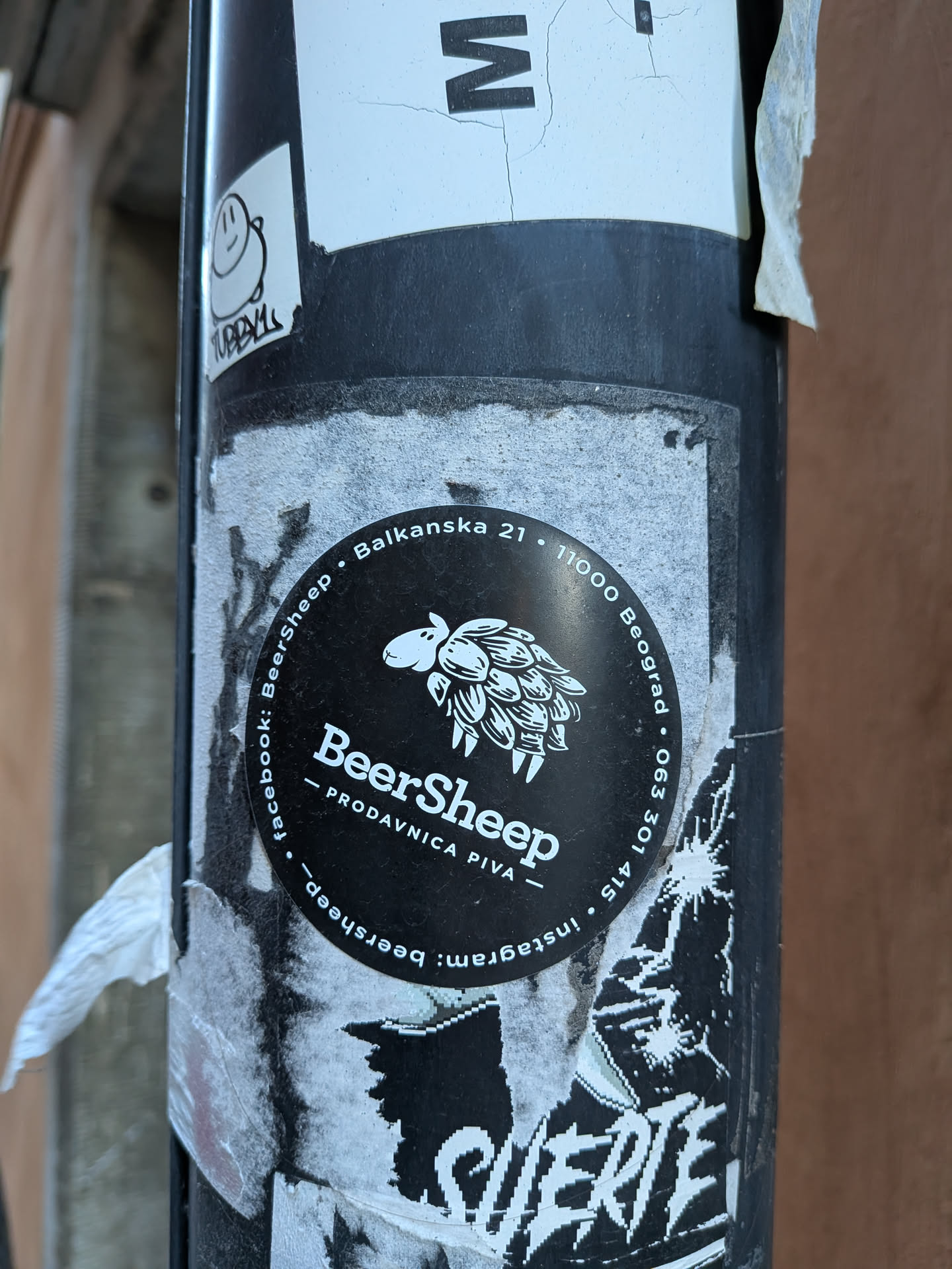 A circular black sticker featuring a white stylized illustration of a sheep whose fleece is composed of hop cones, facing left. Below the illustration, the word 'BeerSheep' is prominently displayed in white, with '— PRODAVNICA PIVA —' (Beer Shop) underneath. Around the perimeter, in white text, are contact details and social media handles: 'Balkanska 21 • 11000 Beograd • 063 301 415 • instagram: beersheep • facebook: BeerSheep'. The sticker has a clean, monochrome design, standing out against the worn pole.