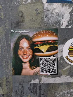 A square sticker featuring a woman with freckles and a big smile, next to a cartoonish burger. Above them is the text 'DJ burgerhead' and below a QR code.
