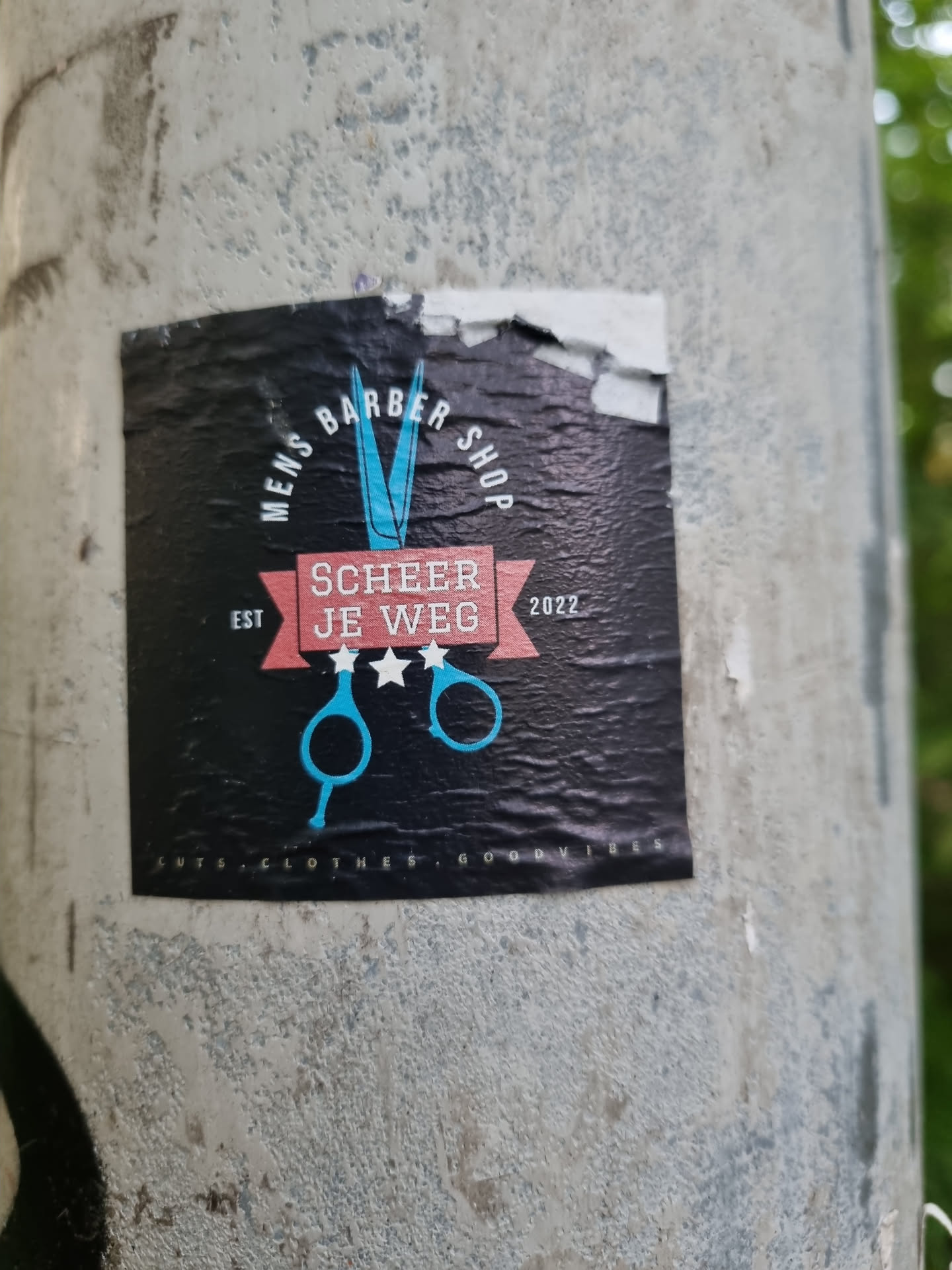Black sticker with a pair of scissors and text. The text includes 'Mens Barber Shop', 'Scheer Je Weg', 'Est 2022', and 'coots, clothes, goodies'.