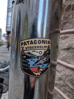 A shield-shaped sticker with a cream-colored border and a thick black outline. The top section features the word 'PATAGONIA' in bold black capital letters, with 'STOCKHOLM' directly below it in a slightly smaller font. To the left of 'STOCKHOLM' is a stylized brown tower with three crenellations, and to its right is a green pine tree. The main body of the sticker depicts a vibrant outdoor landscape: a blue body of water (likely a lake or river) flows through green hills and mountains. A large yellow sun or moon is visible in the sky. On the right bank, a small red cabin with a white roof is nestled among trees. On the left bank, a small white owl-like figure is visible. In the foreground, at the bottom, a red tent with a yellow base is pitched by the water. The overall style is graphic and illustrative, using a palette of greens, blues, reds, yellow, black, and cream.