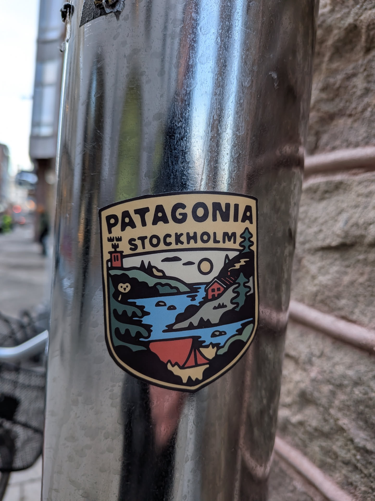A shield-shaped sticker with a cream-colored border and a thick black outline. The top section features the word 'PATAGONIA' in bold black capital letters, with 'STOCKHOLM' directly below it in a slightly smaller font. To the left of 'STOCKHOLM' is a stylized brown tower with three crenellations, and to its right is a green pine tree. The main body of the sticker depicts a vibrant outdoor landscape: a blue body of water (likely a lake or river) flows through green hills and mountains. A large yellow sun or moon is visible in the sky. On the right bank, a small red cabin with a white roof is nestled among trees. On the left bank, a small white owl-like figure is visible. In the foreground, at the bottom, a red tent with a yellow base is pitched by the water. The overall style is graphic and illustrative, using a palette of greens, blues, reds, yellow, black, and cream.