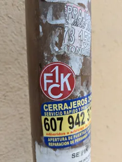 A weathered image shows a pole covered in various stickers. Centered is a red and white round sticker with "FCK" written inside. Below it, a yellow and blue sticker promotes locksmith services, displaying the phone number 607 942 37. The pole also has other partially torn stickers above the "FCK" sticker, with remnants of letters and numbers.