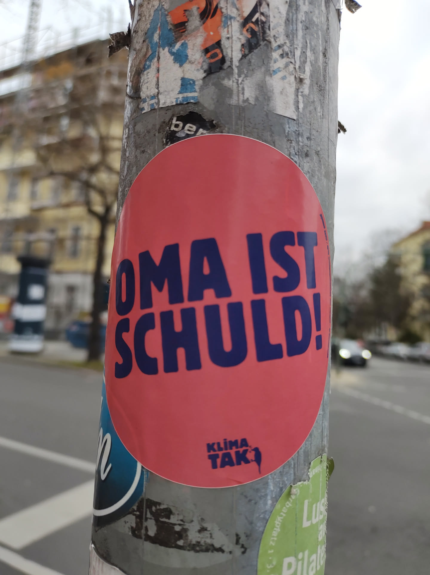 A round pink sticker with the text "OMA IST SCHULD!" in dark blue letters.  At the bottom, a small logo says "Klima Tak". The sticker is affixed to a pole covered in other stickers.