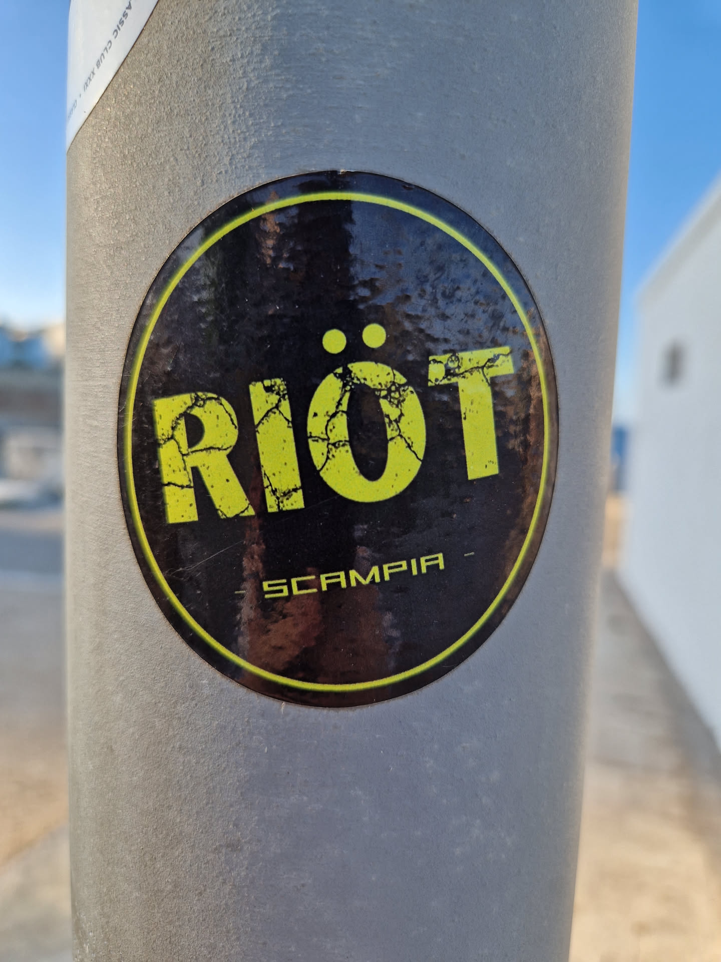 A black and yellow round sticker with the word "RIOT" in a distressed font and "SCAMPIA" written below it. The sticker is affixed to a grey pole.