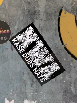 A black and white sticker with the text "KASE DUBS HATS" and a stylized letter M in the center. The letter M is decorated with intricate designs.