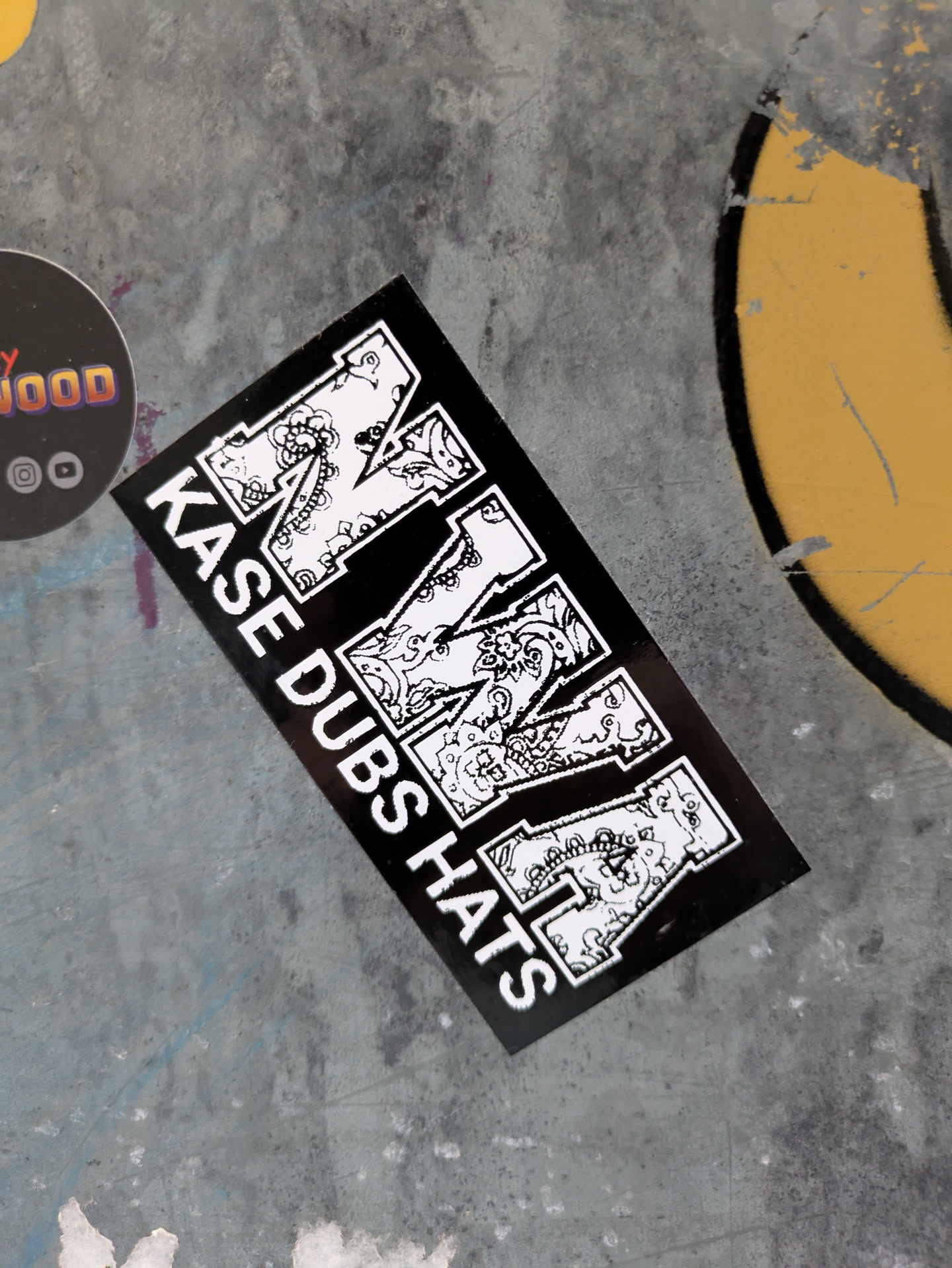 A black and white sticker with the text "KASE DUBS HATS" and a stylized letter M in the center. The letter M is decorated with intricate designs.