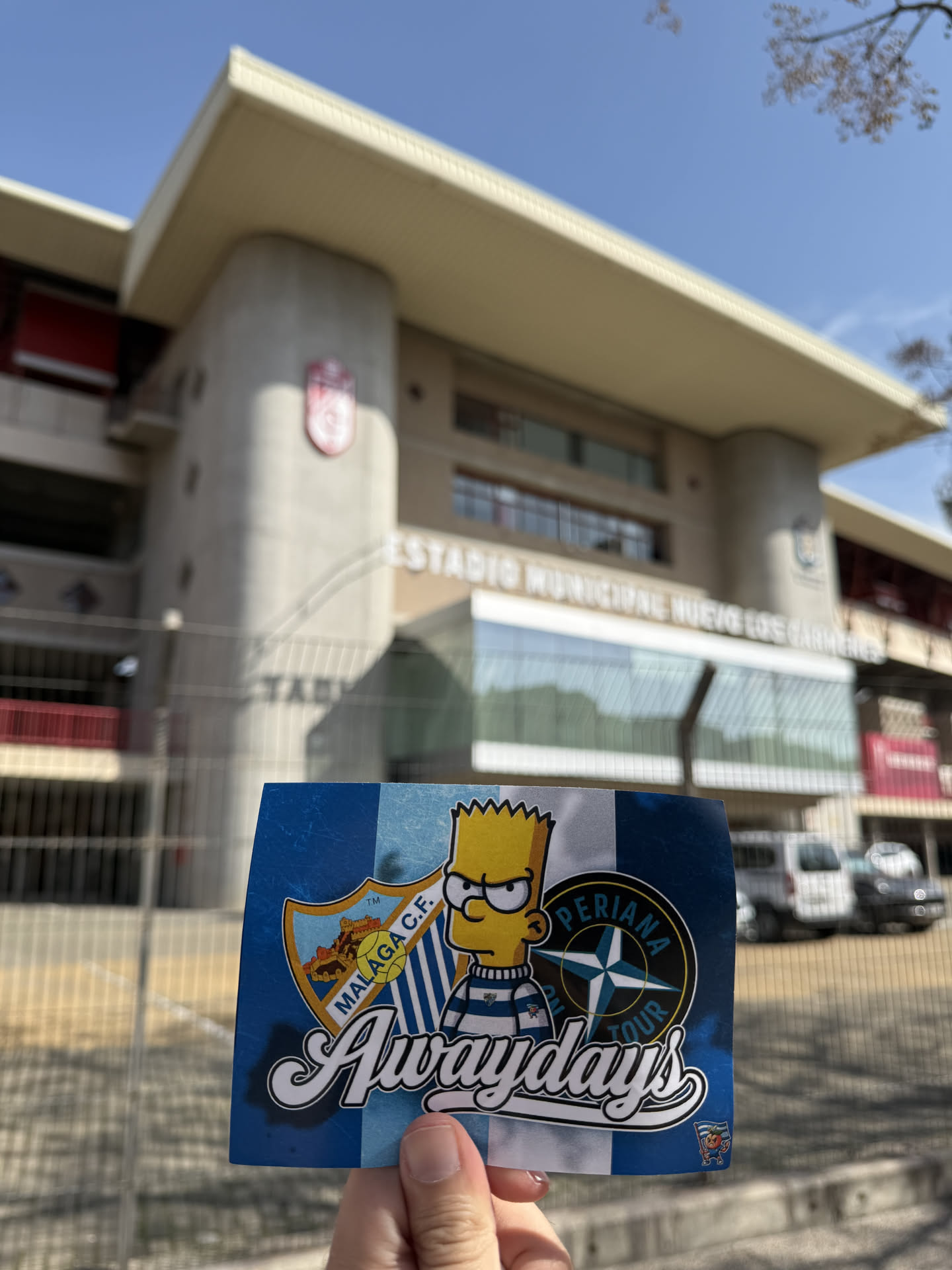 The sticker features a blue and white striped background, reminiscent of a football kit or flag, with a distressed texture. On the left, there is a shield-shaped crest for 'MÁLAGA C.F.' depicting a castle, a sun, and a football. In the center, Bart Simpson is illustrated wearing a blue and white striped shirt and glasses, with a slightly grumpy expression. To his right, a circular emblem with a compass rose design reads 'PERIANA TOUR' around its top edge. The word 'Awaydays' is prominently displayed across the bottom in a large, stylized white script with a black outline. In the bottom right corner, a small additional logo shows a character holding a flag with an orange, with 'PERIANA' written below it. The sticker is held by a hand against a blurred background of a stadium.