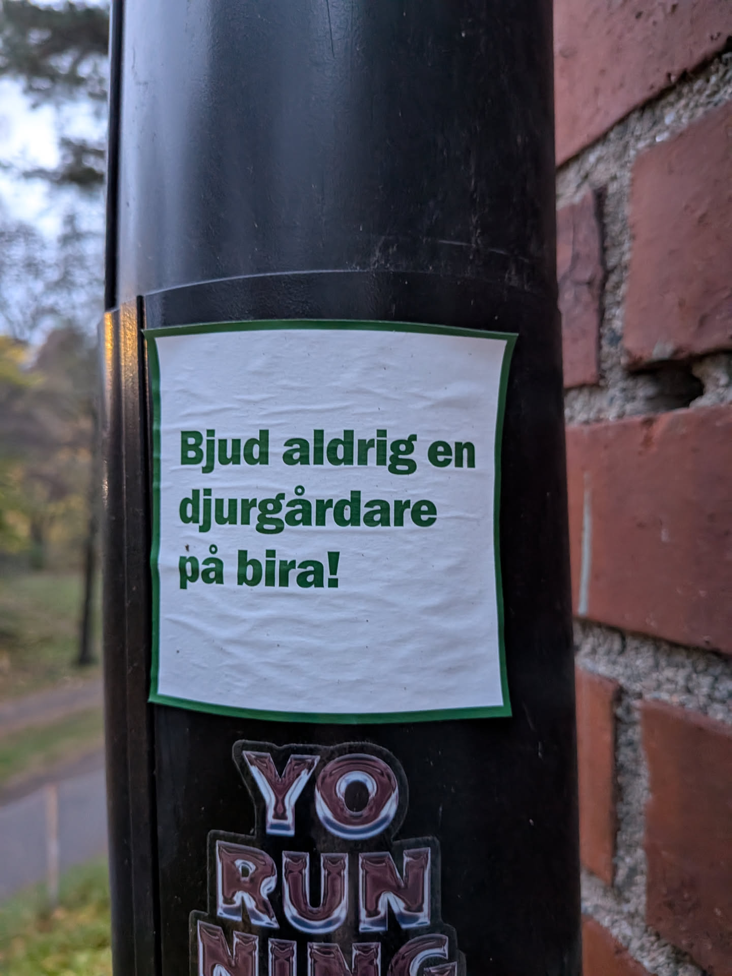The sticker is white with a green border and features text in green capital letters. The text is arranged in three lines. The paper appears slightly creased and textured. Below this sticker, partially visible, are other stickers with metallic-looking lettering that spell out 'YO RUNNING'.
