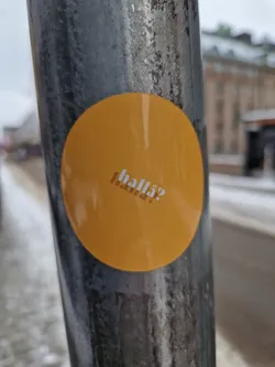 A round, yellow sticker with the word "halla?" in a stylized font. The sticker is affixed to a dark gray metal pole outdoors. The background is slightly blurred, suggesting a city street scene with some snow on the ground.