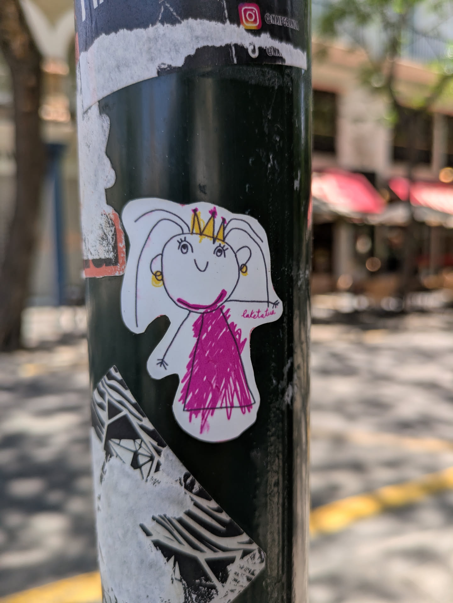 A white sticker of a princess drawn with simple lines, wearing a crown and a pink dress, is adhered to a black metal pole. The drawing has a childish, playful style. There are other stickers above and below it. In the background, a blurry street scene unfolds.