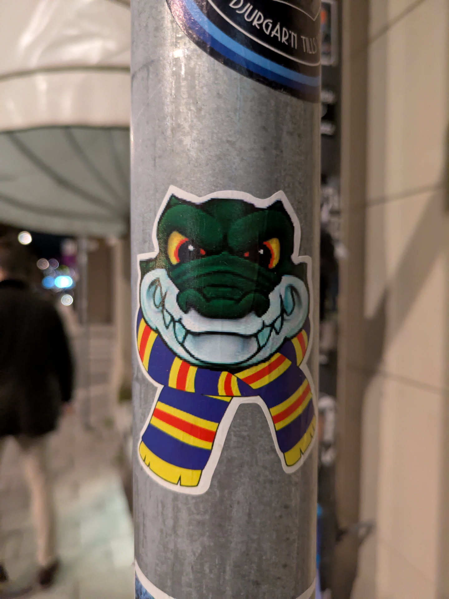 A sticker featuring a grinning alligator wearing a scarf with blue, yellow, and red stripes. The gator has bright yellow eyes and a menacing look.