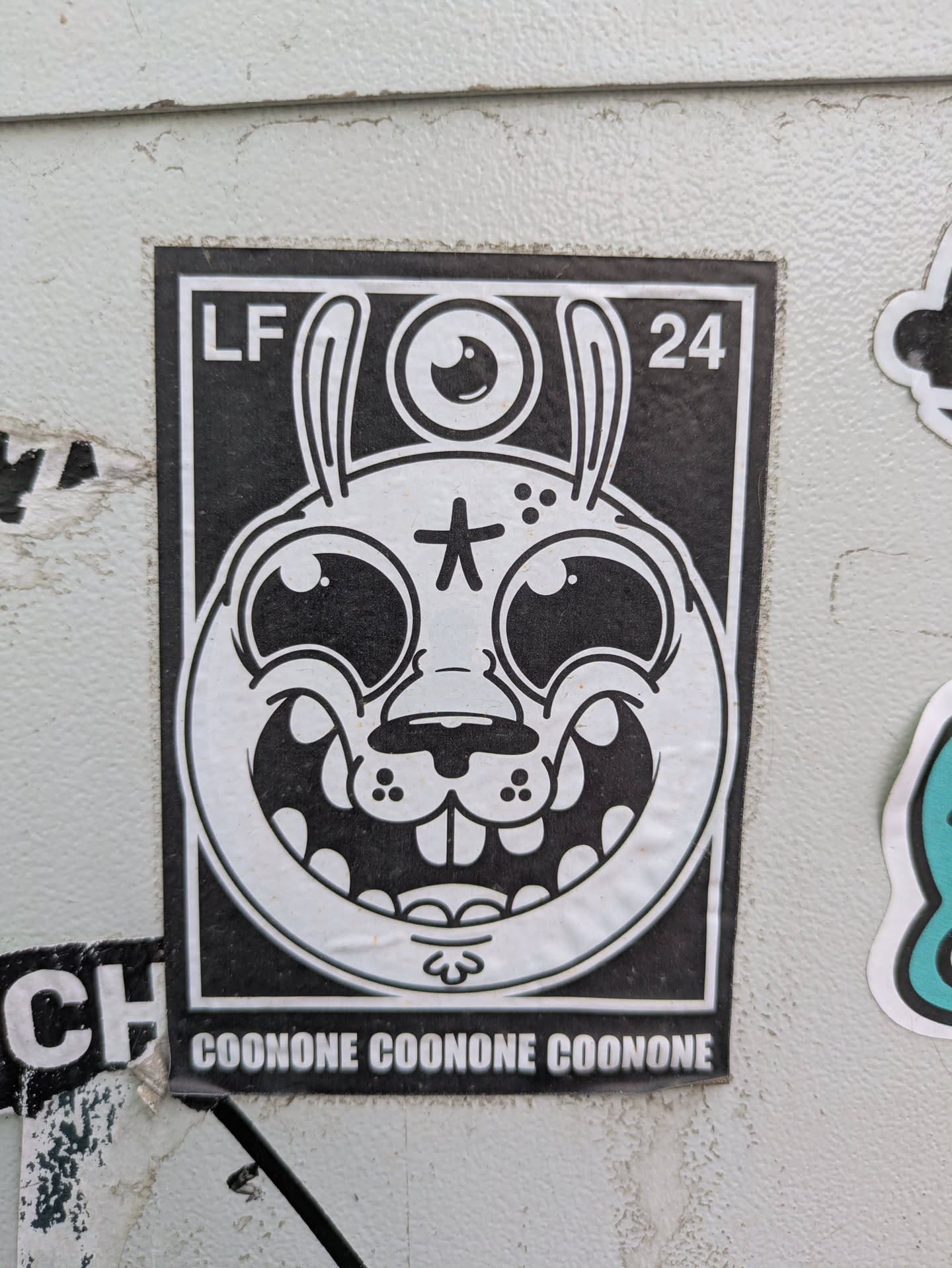 This black and white sticker showcases a cartoonish rabbit-like creature with long ears and a single eye located above its head. The creature has a wide grin, prominent teeth, and a star symbol on its forehead. The letters "LF" and the number "24" are printed in the upper corners, and the word "COONONE" is repeated along the bottom of the sticker.