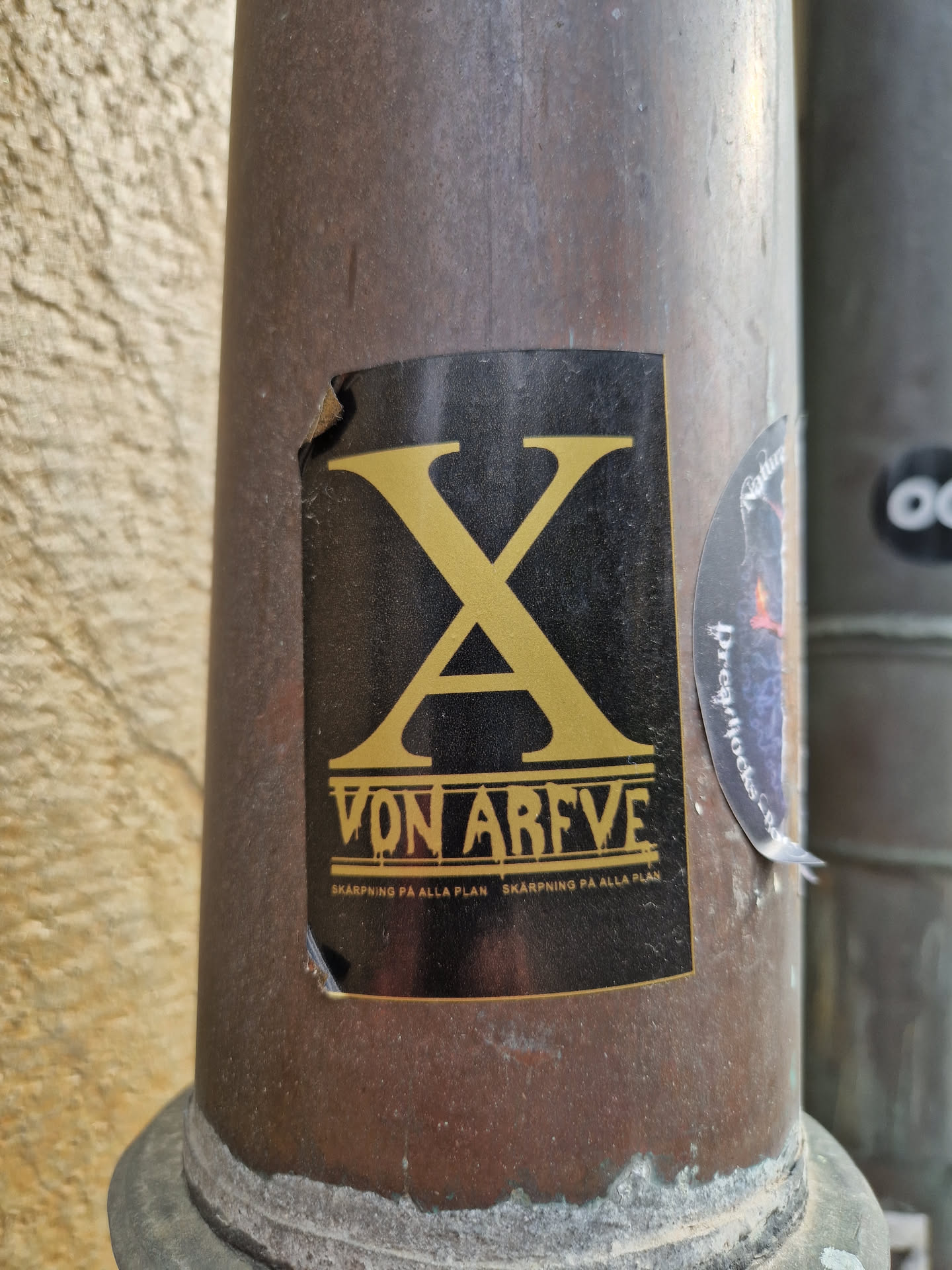 A black sticker with gold lettering on a dark brown pipe. The sticker features a large stylized 'XA' logo, which is likely an abbreviation or a brand logo. Underneath the logo, the text 'VON ARFVE' is displayed, which could be a name, a brand, or a title. Repeated twice below that, there is the text 'SKARPNING PÅ ALLA PLAN' in smaller letters, possibly a slogan or tagline. The sticker has some minor damage and is slightly torn at the top left corner.