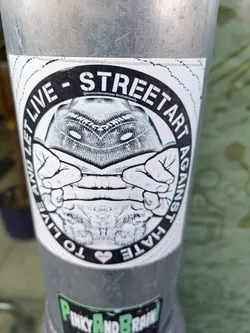 Black and white sticker with an illustration of a person covering their face with their hands. The text around the image says "STREET ART AGAINST HATE - TO LIVE AND LET LIVE". There is a small heart at the bottom of the sticker.  An Instagram handle is visible in small print above the image. 