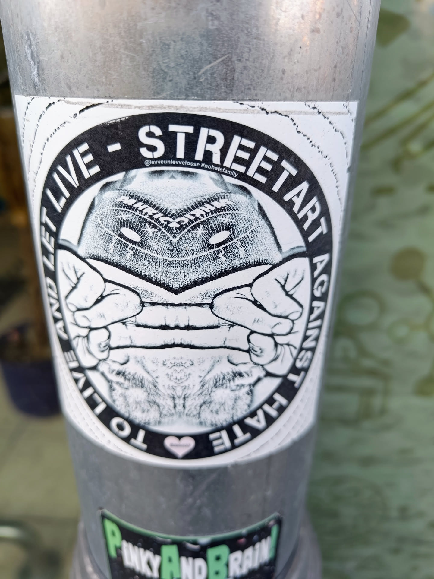 Black and white sticker with an illustration of a person covering their face with their hands. The text around the image says "STREET ART AGAINST HATE - TO LIVE AND LET LIVE". There is a small heart at the bottom of the sticker.  An Instagram handle is visible in small print above the image. 