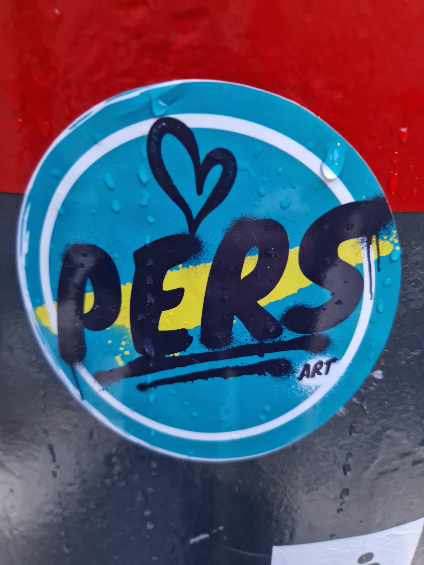 A round sticker with a teal background and the word PERS written in black with yellow accents. There is a small heart above the word. The word ART is written in small letters at the bottom of the sticker.
