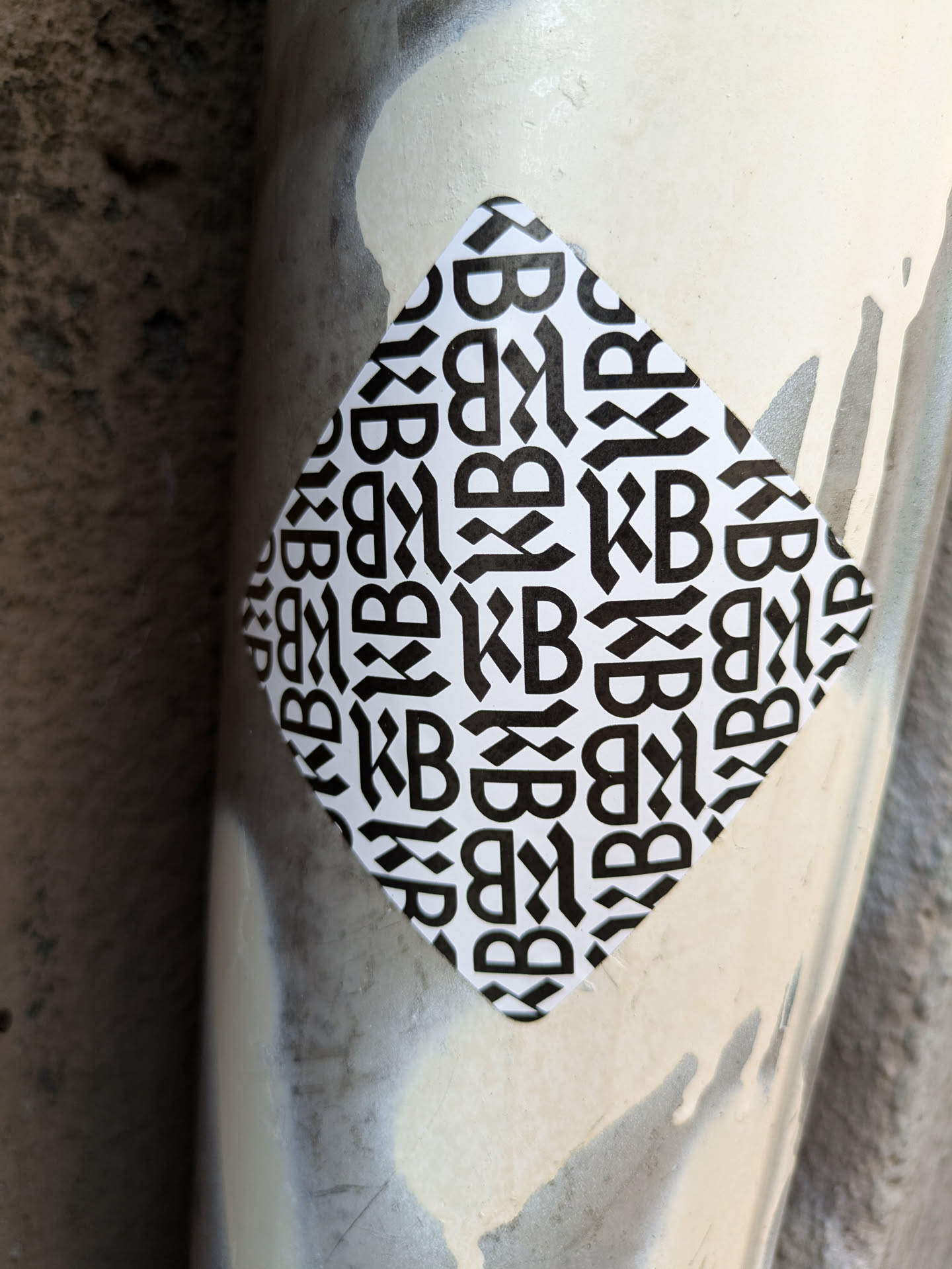 A diamond-shaped sticker with a black and white repeating pattern of the letters KB. The letters are stylized and interlocked within diagonal lines. The sticker is affixed to a metallic surface.