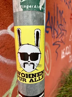 The sticker features a stark white rabbit head with thick black outlines, centered on a bright yellow background. The rabbit wears large, dark black sunglasses, obscuring its eyes, and sports a prominent black handlebar mustache with two small, square front teeth visible beneath its nose. Thin black lines represent whiskers extending from its face. Below the rabbit, bold black uppercase text reads 'WOHNEN FÜR ALLE' (Housing for All) arranged in a slight curve. Along the right edge, in small black vertical text, is an imprint: 'V.i.S.d.P. Hauke Stiewers, Heidestraße 10, 10543 Berlin'. The sticker is applied to a grey metal pole.
