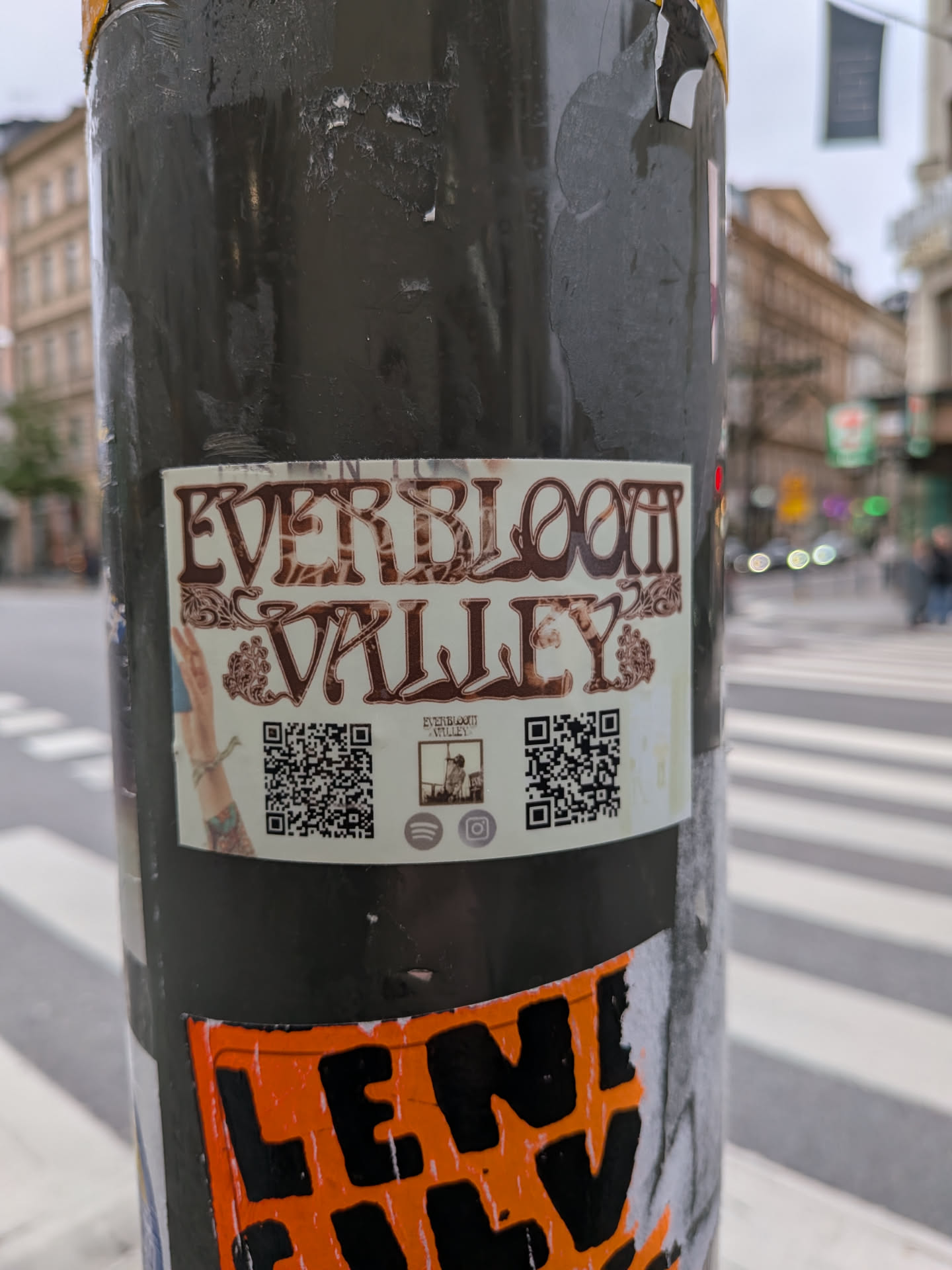 The sticker is rectangular with a light cream background. At the top, the words "EVERBLOOM" are displayed in a large, ornate, brown, serif font with decorative swirls. Below this, "VALLEY" is written in a similar, slightly smaller, ornate brown font. To the left of "VALLEY" is an illustration of a hand with bracelets and tattoos, giving a peace sign. To the right of "VALLEY" are two QR codes. Below the left QR code is a small illustration of a person standing in a landscape, with the text "EVERBLOOM VALLEY" above it. Below the right QR code are icons for Spotify and Instagram.