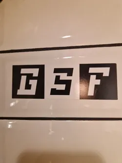 Three black square stickers with white letters GSTF are applied on a white surface. Each letter is on a separate sticker.