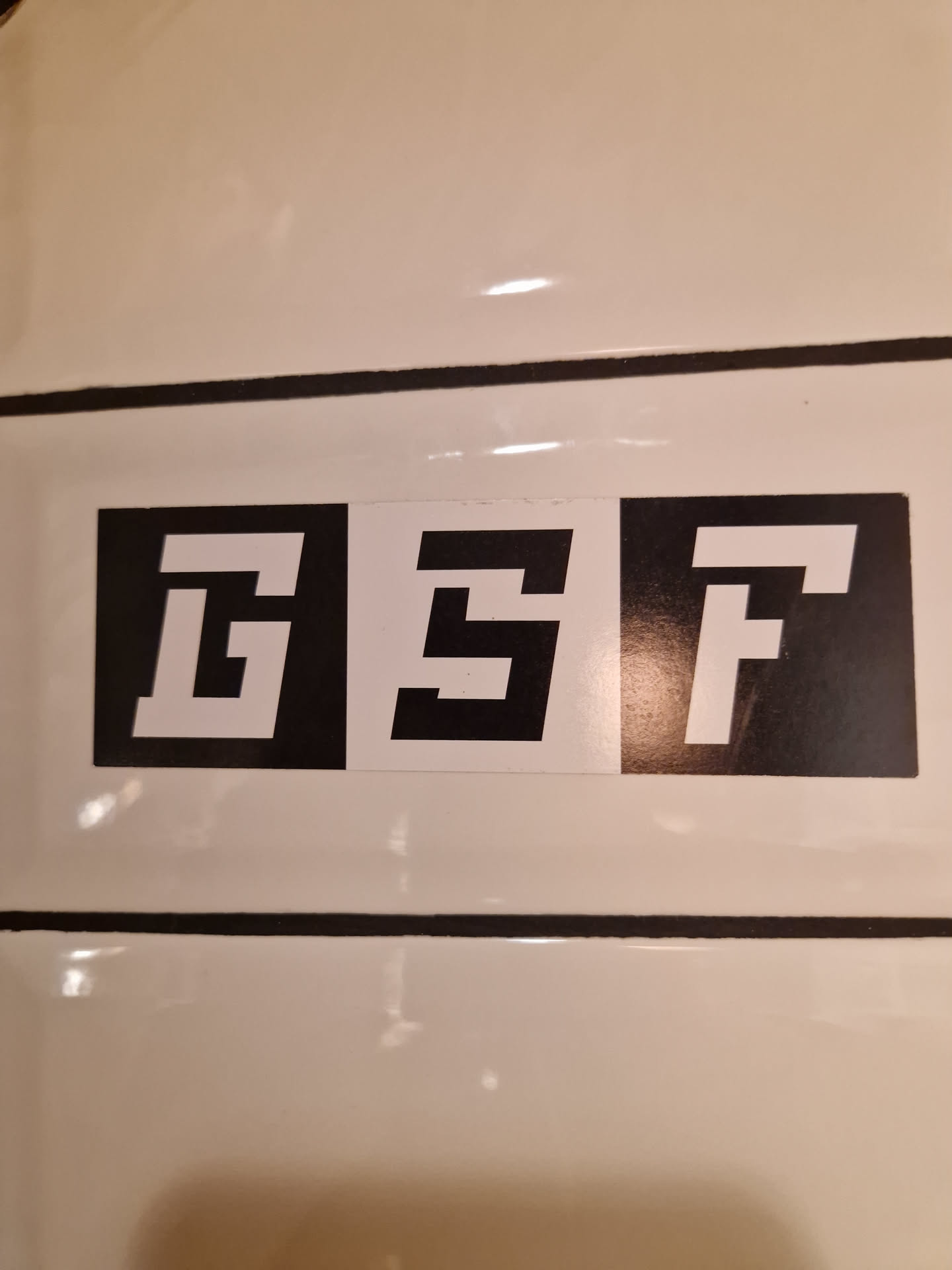 Three black square stickers with white letters GSTF are applied on a white surface. Each letter is on a separate sticker.