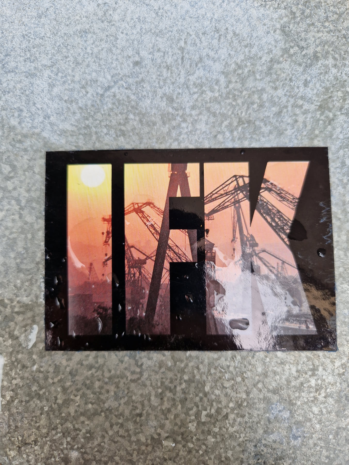 A sticker featuring the letters IFK overlaid on a sunset image of harbor cranes. The sticker has a slightly worn look, suggesting it may have been placed outdoors for some time. 