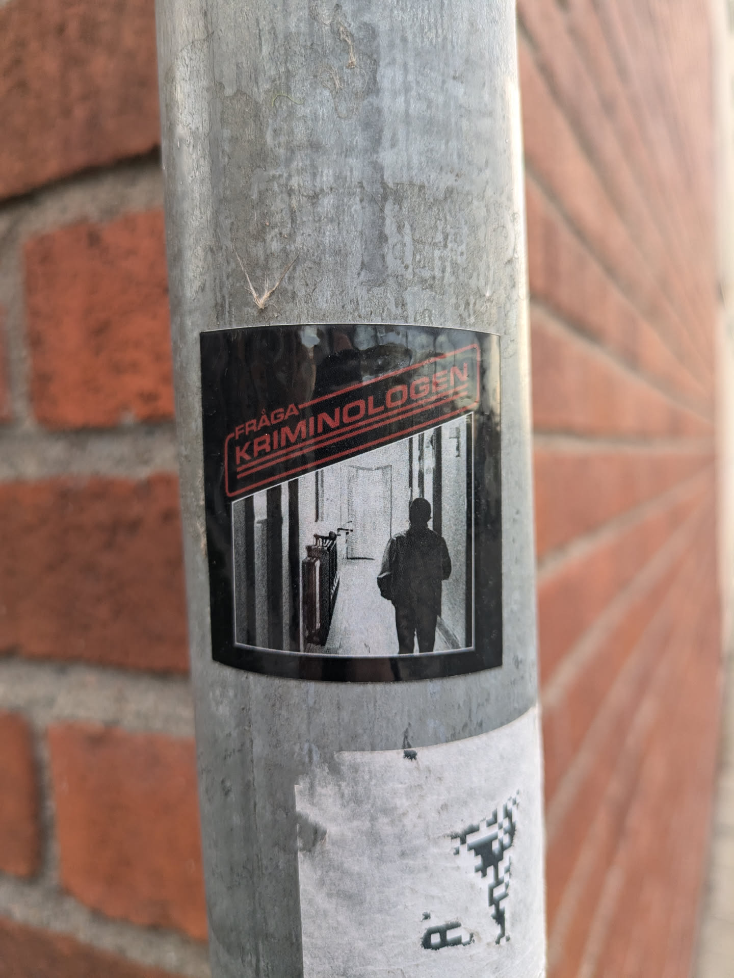 A black and white sticker featuring a person walking down a hallway with the text "Fråga Kriminologen" (Ask the Criminologist) in red and black. The sticker is affixed to a metal pole against a brick wall.
